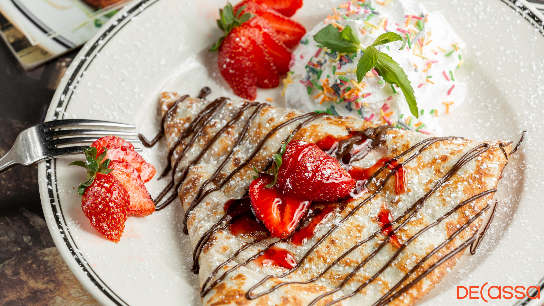 A beautiful Crepe Pancake on a white plate with chocolate sauce and topped with strawberries with a side of whipped cream