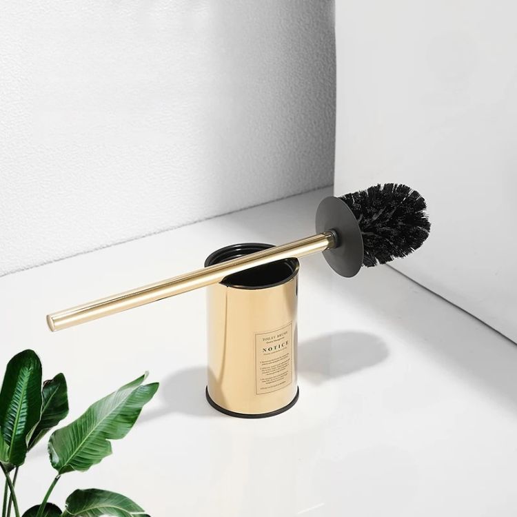 Gold toilet brush holder with brush placed on floor.