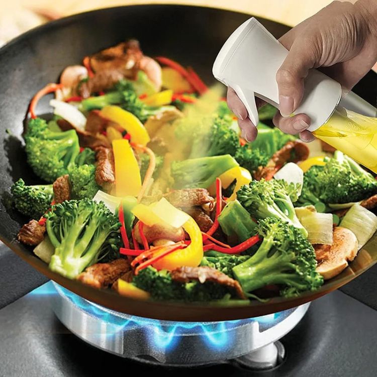 Hand spraying oil over colorful vegetables in a wok using an oil sprayer bottle.
