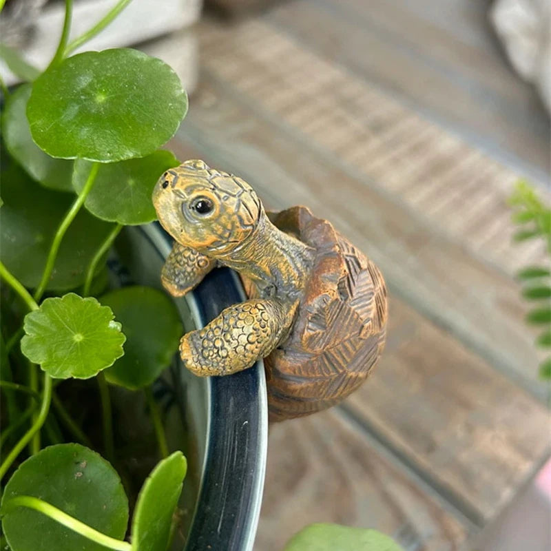 Realistic turtle garden sculpture hanging on a pot rim with green leaves in the background.