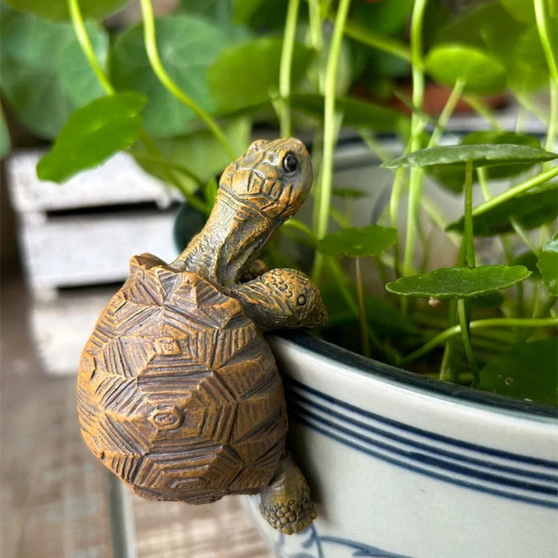 Turtle garden sculpture climbing the edge of a ceramic plant pot surrounded by greenery.
